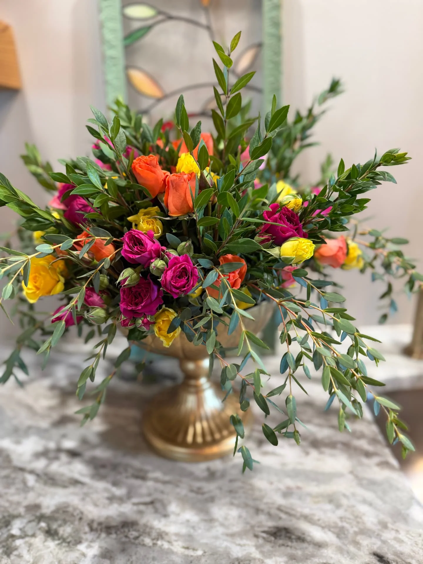 Bright orange, yellow and fuchsia roses with trailing greenery in a gold pedestal urn